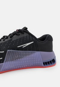 Chaussure de sport noire avec une tige en mesh texturée, une semelle intermédiaire violette et une semelle extérieure rouge, vue de côté avec lacets sur fond blanc.