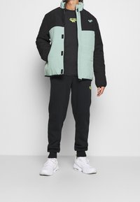 Doudoune vert clair et noir avec un col haut, portée sur un t-shirt noir avec un logo vert fluo, associée à un jogging noir et des baskets blanches.