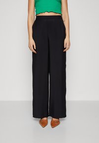 Pantalons larges noirs en tissu texturé, dotés d'une taille haute et d'une coupe décontractée. Associés à des ballerines brunes à bouts pointus.