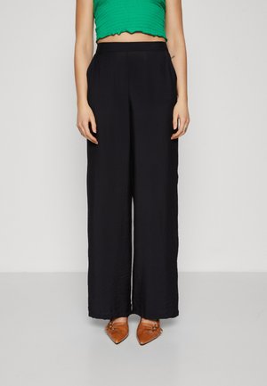 Pantalons larges noirs en tissu texturé, dotés d'une taille haute et d'une coupe décontractée. Associés à des ballerines brunes à bouts pointus.