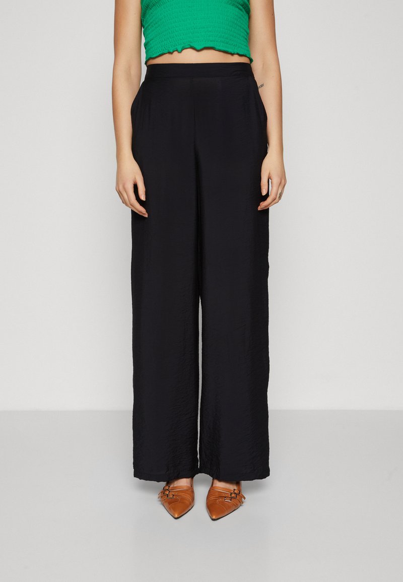 Pantalons larges noirs en tissu texturé, dotés d'une taille haute et d'une coupe décontractée. Associés à des ballerines brunes à bouts pointus.