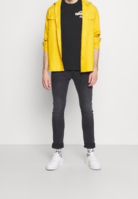 Žlutá košile na knoflíky přes černé tričko, kombinovaná s tmavými džínami a bílými teniskami. Na sobě má vzorované ponožky a vyhrnuté nohavice.