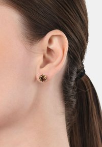 Pendientes de botón redondos con una gema marrón de superficie facetada, montados en un sencillo marco de metal, llevados en la oreja de un modelo.