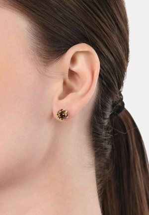 Orecchini a cerchio con una pietra preziosa marrone dalla superficie sfaccettata, incastonati in una semplice montatura metallica, indossati all'orecchio di un modello.