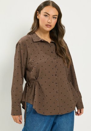 Frau trägt eine braune Bluse mit Knöpfen und schwarzen Punkten sowie seitlichen Kordeln, kombiniert mit blauen Jeans, sie steht vor einem schlichten Hintergrund.