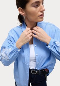 Camicia azzurra a righe con dettagli floreali ricamati sulle maniche, indossata sopra una maglietta bianca, abbinata a una cintura nera e jeans scuri.