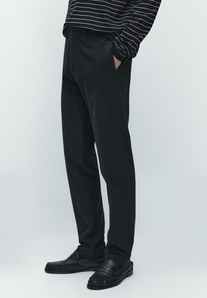 Osoba oblečená v černých rovně střižených kalhotách, černých mokasínách a černé košili s bílými vodorovnými pruhy na dlouhý rukáv, ruce v kapse.