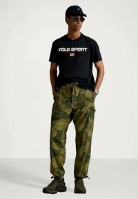 Mężczyzna w czarnej koszulce polo marki Polo Sport, w kamuflażowych spodniach cargo, czarnych sneakersach, czapce i okularach przeciwsłonecznych, stojący z rękami w kieszeniach.