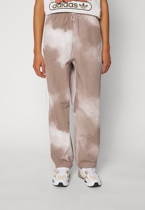 Personne portant un pantalon de survêtement Adidas tie-dye beige et blanc avec des baskets blanches et beiges, debout devant un fond uni.