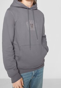 Felpa grigia con cappuccio e tasca frontale, caratterizzata da un design a stemma ricamato. Realizzata in materiale morbido con polsini e orlo a costine.