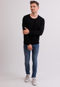 Schwarzer gestrickter Pullover mit rundem Halsausschnitt, kombiniert mit eng anliegenden blauen Jeans und weißen Sneakers. Einfaches Design und glatte Textur.