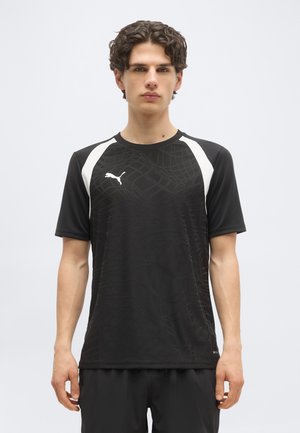 Νεαρός άνδρας που φοράει μαύρο αθλητικό μπλουζάκι Puma με διακριτικό υφασμάτινο μοτίβο και λευκές λεπτομέρειες στους ώμους, όρθιος μπροστά σε απλό φόντο.