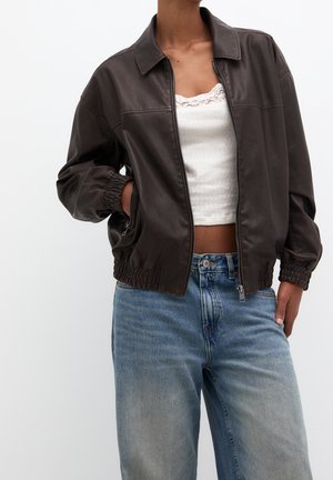 Personne portant une veste en cuir marron foncé zippée sur un débardeur blanc orné de dentelle, associée à un jean denim bleu large, une main dans la poche.