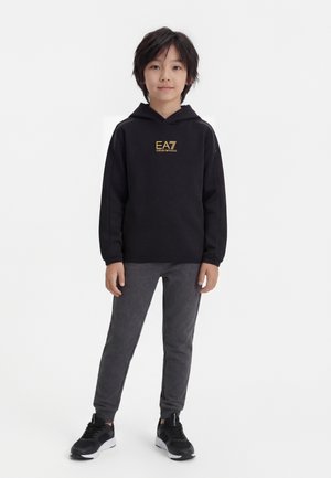 Laps seisab, kandes musta kapuutsiga pusa kullakarva "EA7 Emporio Armani" logoga, tumedat halli pükse ja musti tossusid valgel taustal.