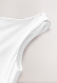 Tissu en coton blanc à la texture lisse, présentant un col arrondi et des manches courtes. Les détails de couture sont minimaux et discrets.