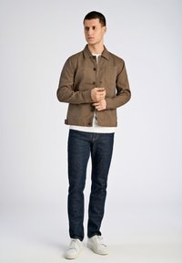 Brun canvasjacka med krage, framknäppning och bröstficka. Bärs över en vit skjorta och matchas med mörka jeans och vita skor.