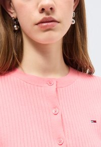 Νεαρή γυναίκα που φοράει ασημένια κρίκους σκουλαρίκια με στρογγυλά κρεμαστά και ένα ριμπ πουλόβερ ροζ με κουμπιά, διακοσμημένο με ένα μικρό κόκκινο, λευκό και μπλε λογότυπο.