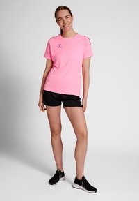 Camiseta deportiva rosa de manga corta, con logo en el pecho, combinada con pantalones cortos negros y zapatillas negras. Tejido suave, diseño ajustado.