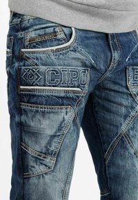 Jean en denim bleu avec plusieurs poches, des coutures complexes et un logo brodé sur le devant. Tissu texturé avec un aspect usé.