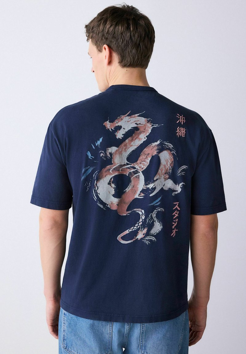 Mees näoga eemale, kandes tumesinist T-särki, mille taga on suur stiliseeritud draakoni kujutis ja Jaapani tekst, ning hele sinised teksad.