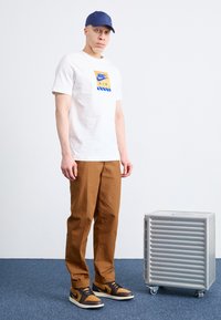 Balts kokvilnas T-krekls ar zilo un dzelteno Nike logo, brūni brīvi bikses, melni un brūni sporta apavi un tumši zils cepure. Blakus ir sudraba glabāšanas vienība.