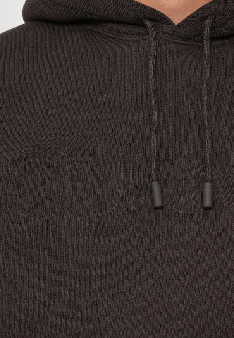 Felpa con cappuccio marrone scuro con una finitura testurizzata, caratterizzata da un logo discreto in rilievo sulla parte frontale e cordini con punte scure.