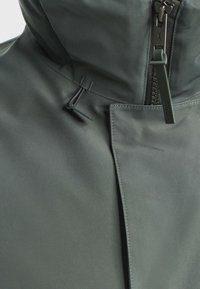 Vodoodolná bunda v tmavosivej farbe, s vysokým golierom, zipsovým uzáverom a nastaviteľným stiahnutím. Hladká textúra s minimálnymi akcentmi.