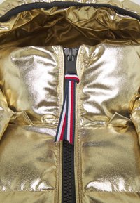 Detailní záběr lesklé zlaté prošívané bundy s černým zipem a táhlem na zipu z tkaniny v červené, bílé a námořnické pruhované barvě.