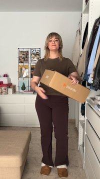 Brūns Zalando kaste turēta rokā, uzvilkts piegulošs brūns krekls, tumši brūnas bikses un brūni čības ar baltu odere. Minimalistisks fons.
