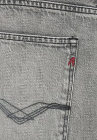 Γκρι ύφασμα τζιν με ορατή πίσω τσέπη. Διαθέτει λεπτομέρεια με κόκκινη κεντητή κορδέλα και μαύρη ραφή κατά μήκος της άκρης της τσέπης.