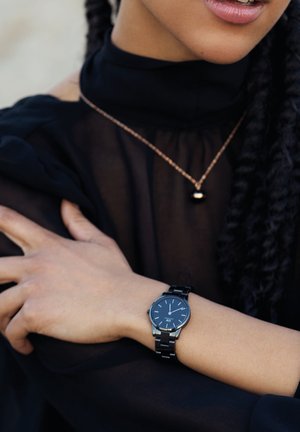 Montre en métal noir avec un cadran rond, index en argent, et un bracelet assorti, porté sur un poignet nu contre un haut transparent noir.