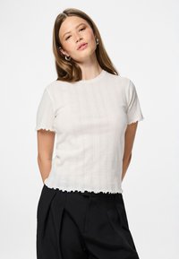 Mlada ženska z dolgimi lasmi nosi belo kratkosleeveno pleteno majico z valovitim robom in črne plisirane hlače, stoji ob belem ozadju.