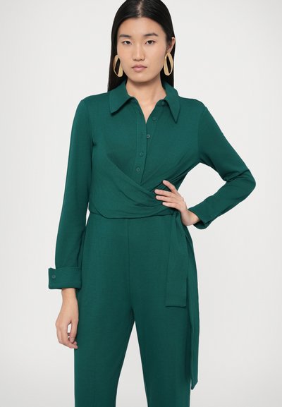 Zöld deréknál átkötős jumpsuit hosszú ujjakkal, gombos elejű és övvel ellátva. Simagázású anyag, inggallérral és mandzsettás ujjakkal.