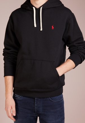 Mężczyzna w czarnej bluzie z białymi sznurkami i czerwonym logo, ręka w przedniej kieszeni, w zestawieniu z ciemnoniebieskimi dżinsami.