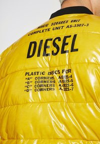Giacca imbottita gialla con finitura lucida e trapuntata. Presenta grandi scritte nere "DIESEL" e dettagli stampati su dischi di plastica e angoli.