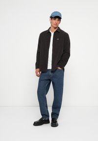 Chemise en velours côtelé marron, t-shirt blanc, casquette bleue, jean bleu et chaussures noires. La chemise a une poche et un petit logo. Coupe décontractée et légère.