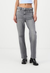 Naissoasu mustade kontsakingade, kõrge vöökohaga hallide teksade ja lühikese helesinise topiga, seismas ühtlase valge tausta ees.