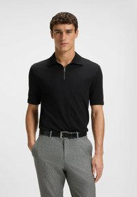 Czarna koszulka polo z krótkim zamkiem, z miękkiego materiału i płaskim kołnierzykiem, zestawiona z szarymi spodniami w wzory i czarnym paskiem.