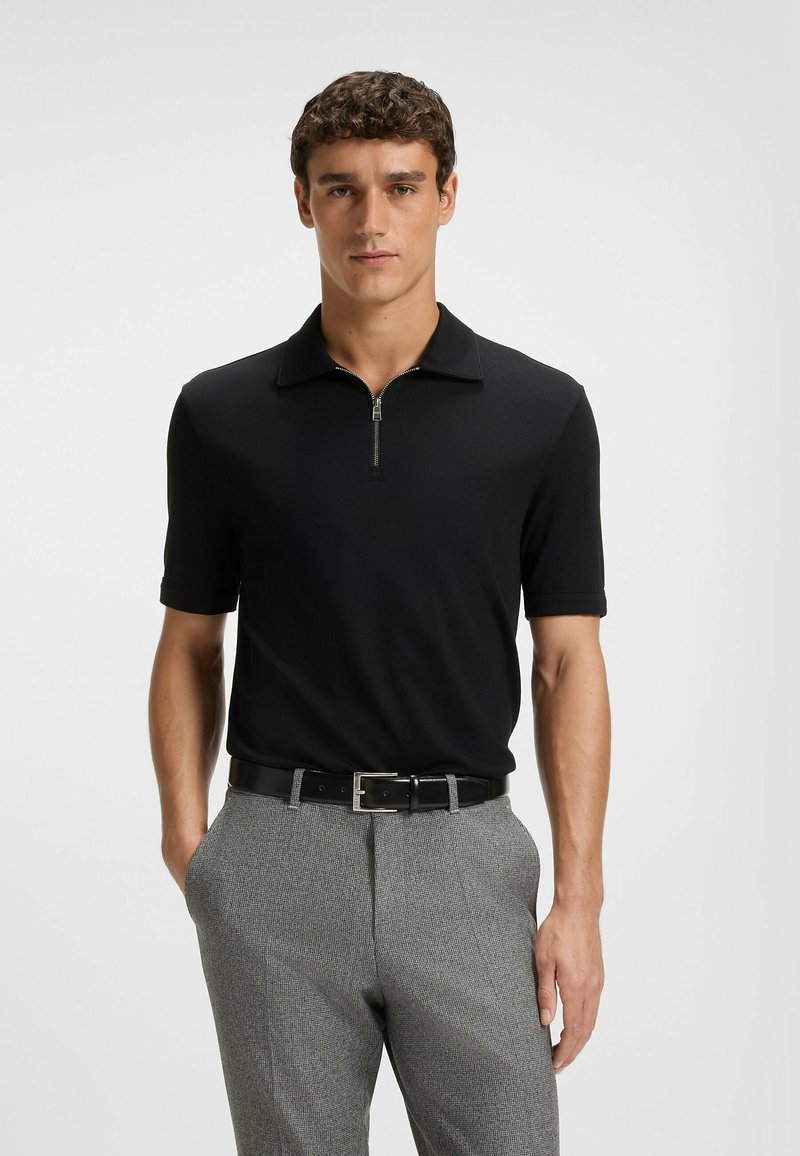 Czarna koszulka polo z krótkim zamkiem, z miękkiego materiału i płaskim kołnierzykiem, zestawiona z szarymi spodniami w wzory i czarnym paskiem.