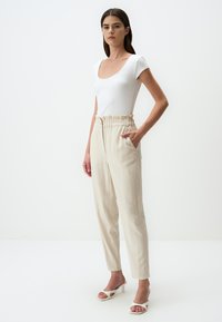 Haut blanc côtelé ajusté associé à un pantalon beige taille haute avec une taille à cordon et des poches latérales. Modèle portant des sandales à talons blanches.