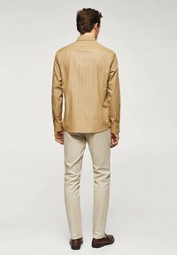 Camicia a maniche lunghe, marrone chiaro con righe verticali, abbinata a pantaloni color crema e scarpe marrone scuro, vista da dietro.