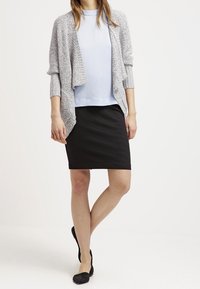 Cardigan grigio lavorato a maglia sopra una maglietta azzurra chiaro, abbinato a una gonna a tubino nera e ballerine nere. L'outfit presenta una vestibilità rilassata e texture morbide.