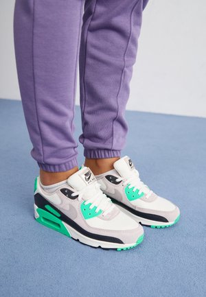 Osoba nosząca białe, czarne i zielone buty Nike Air Max z białymi sznurowadłami oraz fioletowe spodnie dresowe, stojąca na niebieskim dywanie.