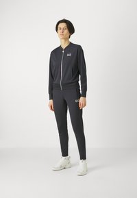 Čierny športový teplákový súbor vyrobený z mäkkej tkaniny, ktorý obsahuje zipsovú bundu a priliehavé nohavice. Výrazný logový detail na hrudi a stehne.