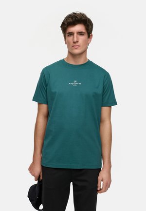 Teal bomulls T-skjorte med korte ermer, rund hals, og hvit logo designet på midten av fronten, kombinert med mørke bukser og en caps.