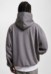 Overdimensjonert grå hettegenser med stor hette, senkede skuldre og ribbestrikket kant. Glatt tekstur og minimalistisk design, båret over en hvit skjorte.