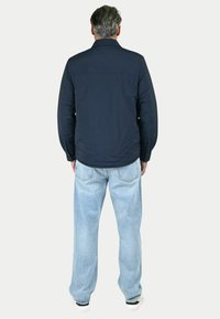 Giacca navy con una trama liscia e colletto a bottoni, abbinata a jeans chiari a gamba larga e sneakers bianche, vista da dietro.