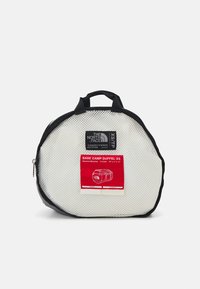 Sac de sport rond en mesh blanc avec des accents noirs, présentant une étiquette rouge. Fermeture zippée et doubles poignées supérieures pour un transport facile.