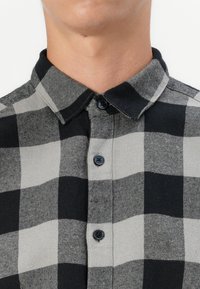 Camisa de cuadros negra y gris con cuello abotonado, dos botones oscuros y patrones a cuadros en contraste de diferentes tamaños.