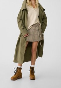 Trench coat verde oliva sopra un maglione beige a scollo a V, abbinato a una mini gonna a quadri e stivali marroni con lacci e suola spessa.